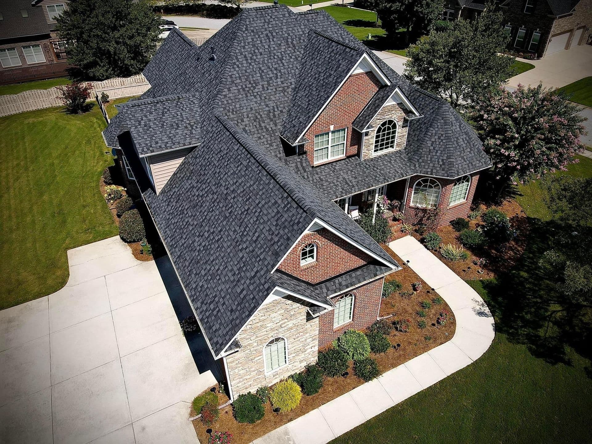An aerial view of a brick and stone house with newly-installed gray asphalt shingle roofing.